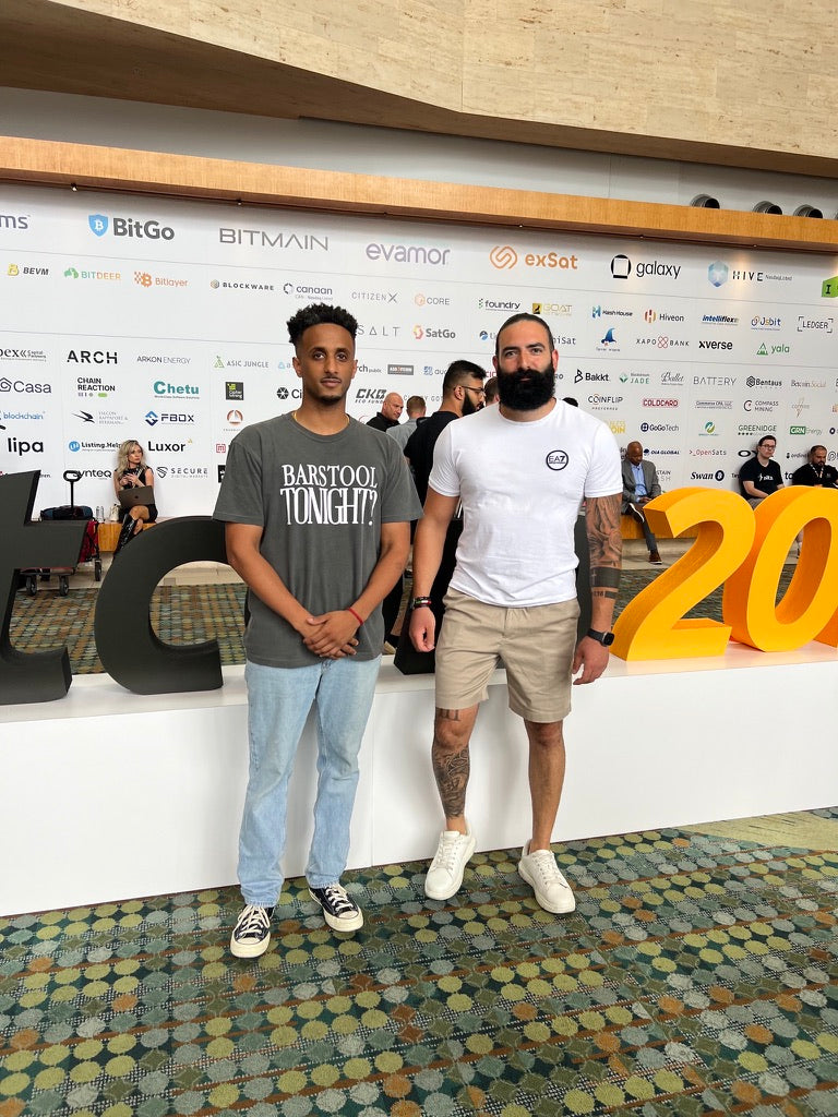 Two founders at a Bitcoin industry conference, in front of the sponsor wall